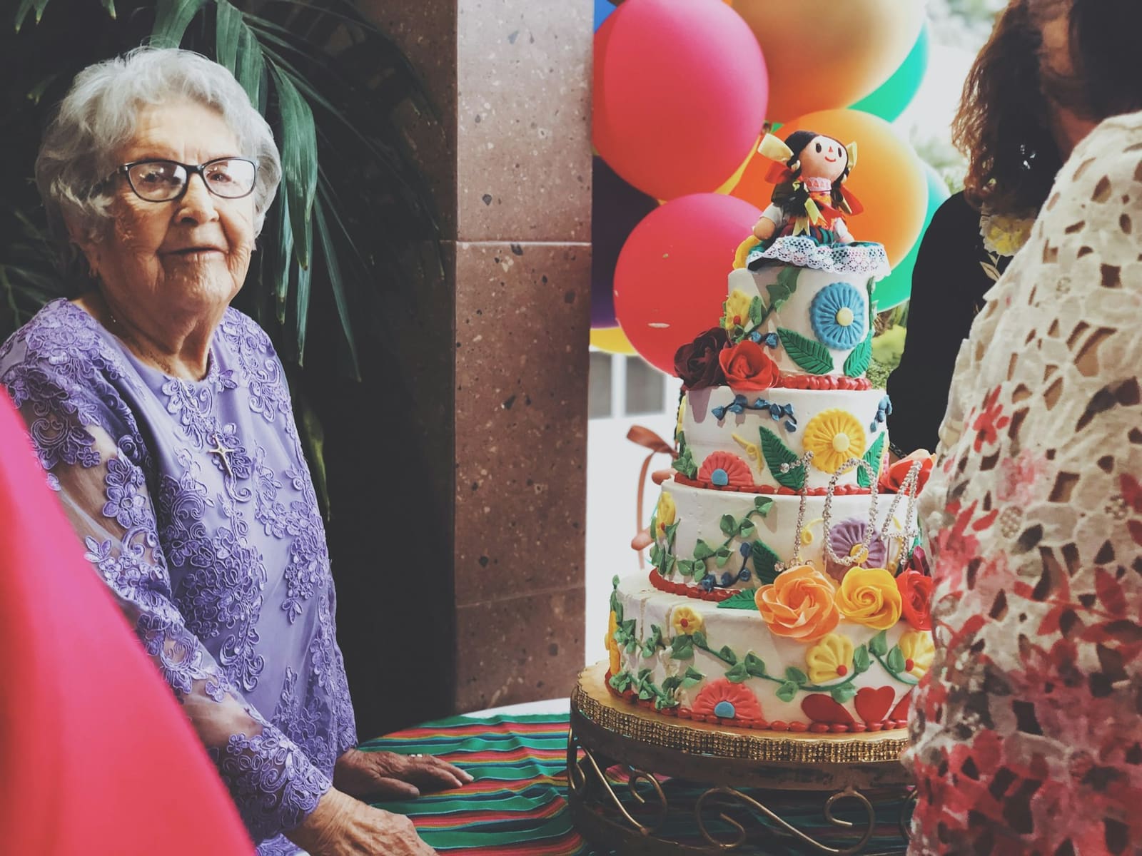 An older woman smiling beside a colourful birthday cake surrounded by balloons